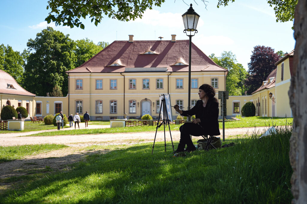 Eine Künstlerin malt auf einer Staffelei vor einem kleinen, gelben Barockschloss