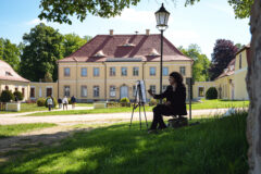 Eine Künstlerin malt auf einer Staffelei vor einem kleinen, gelben Barockschloss