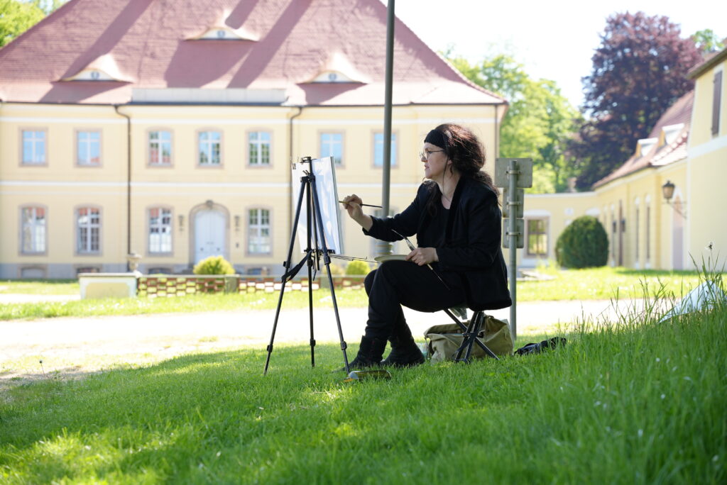 Eine Künstlerin malt auf einer Staffelei, im Hintergrund ein kleines Barockschloss